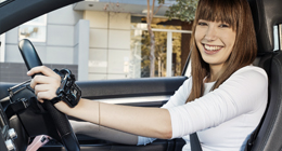 A cheerful woman in a white shirt is behind the wheel of a car.